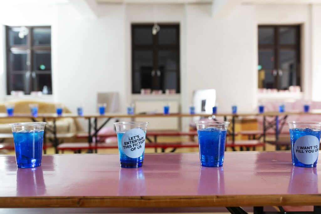 cups with very bright blue drinks the have written "lets entertains this fiction of us" and another cup with "I want to fill you up"
HAUT YC Festival 2023